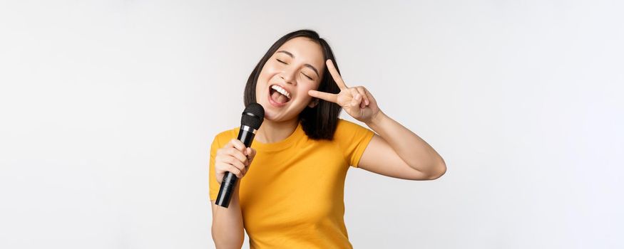 Happy asian girl dancing and singing karaoke, holding microphone in hand, having fun, standing over white background.