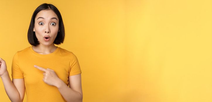 Smiling asian woman pointing fingers left, showing advertisement on empty copy space, standing over yellow background.