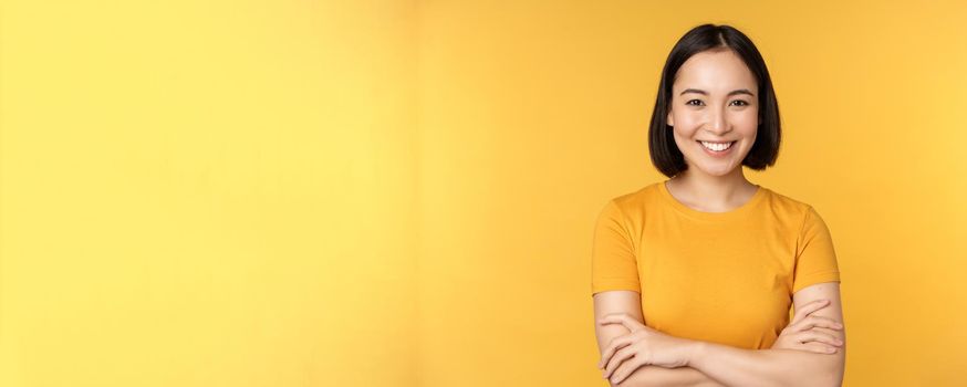 Confident asian girl cross arms on chest, smiling and looking assertive, standing over yellow background.