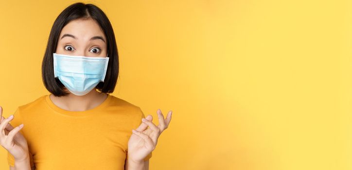 Covid-19 and medical concept. Close up portrait of asian woman in face mask, looking surprised and amazed at news, standing over yellow background.