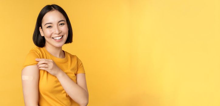 Vaccination and covid-19 pandemic concept. Happy and healthy asian girl pointing at her shoulder with band aid after vaccinating from coronavirus, yellow background.