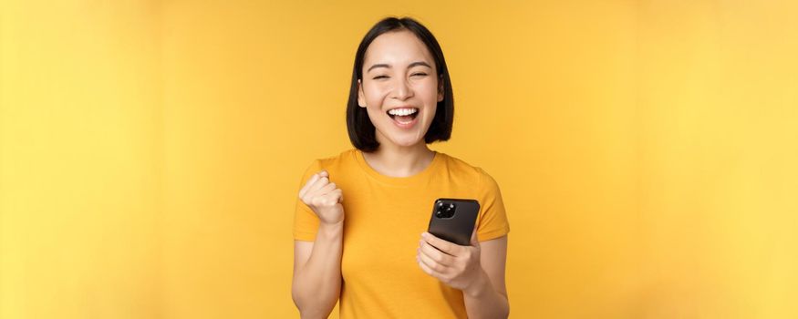 Joyful asian woman celebrating, holding mobile phone, winning, achieve goal on smartphone, standing over yellow background.