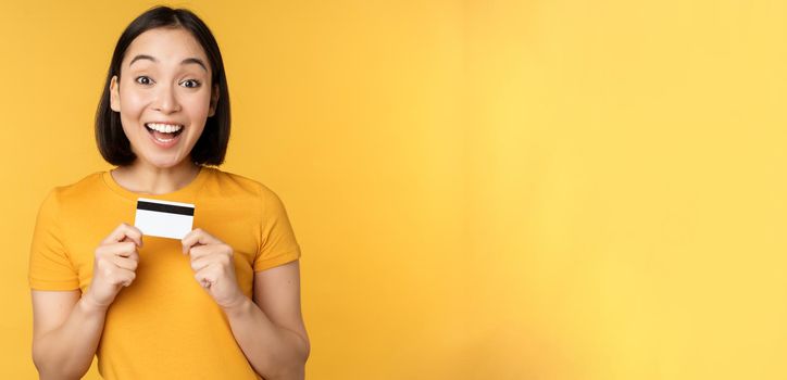 Beautiful asian woman showing credit card and smiling, recommending bank service, standing over yellow background. Copy space