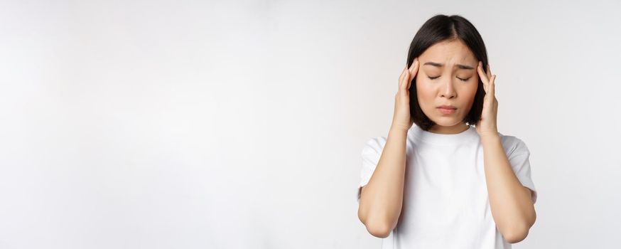 Portrait of asian girl feeling headache, migraine or being ill, standing in white t-shirt over white background. Copy space