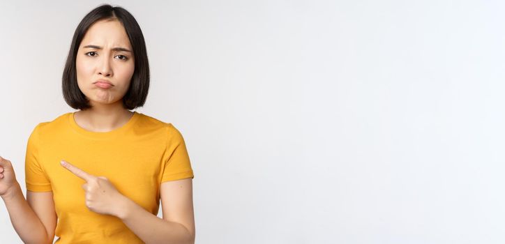 Portrait of disappointed brunette asian girl, pointing fingers left, grimacing upset, showing smth unpleasant, standing over white background.