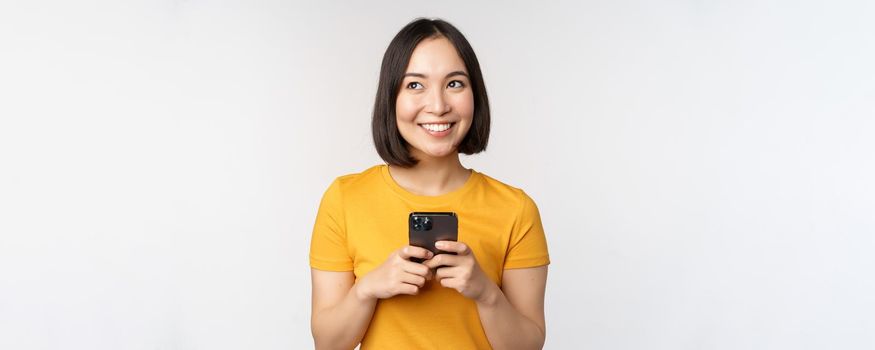 People and technology concept. Smiling asian girl using smartphone, texting on mobile phone, standing against white background.