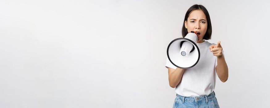 Angry asian woman with megaphone, scolding, accusing someone, protesting with speakerphone on protest, standing over white background.