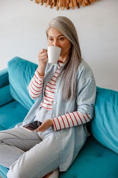 Female person holding smartphone and drinking hot beverage while sitting on couch in her apartment