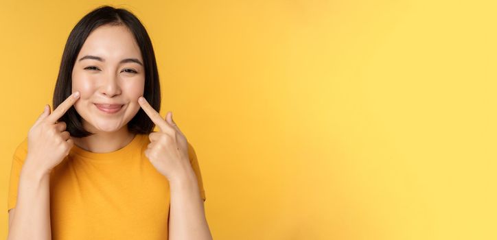 Close up portrait of cute asian girl showing her dimples and smiling coquettish at camera, standing over yellow background. Copy space