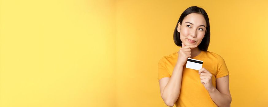 Thinking asian woman holding credit card and smiling, planning buy smth, standing thoughtful over yellow background.