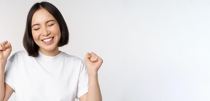 Dancing asian girl celebrating, feeling happy and upbeat, smiling broadly, standing over studio white background.