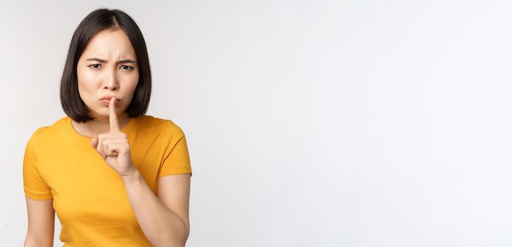 Portrait of angry asian woman shushing, press finger to lips, taboo stop talking sig, looking annoyed, scolding, standing over white background.