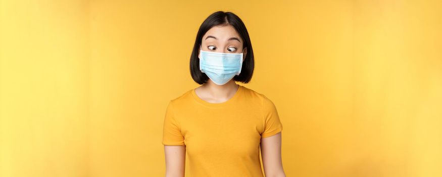 Health and people concept. Funny asian girl squinting, looking at her face medical mask, standing over yellow background.