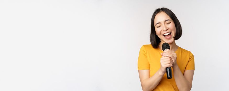 Happy asian girl singing and having fun, holding microphone at karaoke, standing in yellow tshirt against white background. Copy space