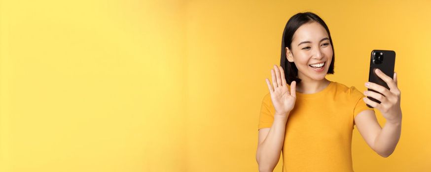 Image of happy, beautiful asian girl video chatting, talking on smartphone application, standing against yellow background. Copy space