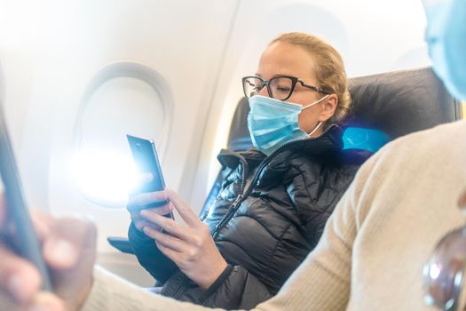 A young couple wearing face mask, using smart phone while traveling on airplane. New normal travel after covid-19 pandemic concept