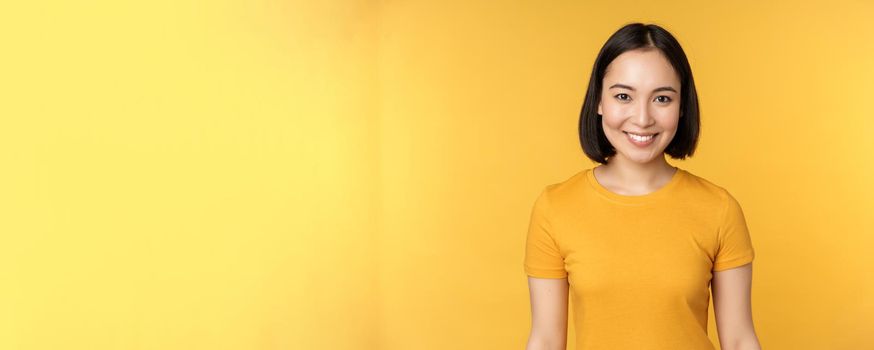 Portrait of young modern asian woman, smiling happy with white teeth, looking confident at camera, wearing casual t-shirt, standing over yellow background.