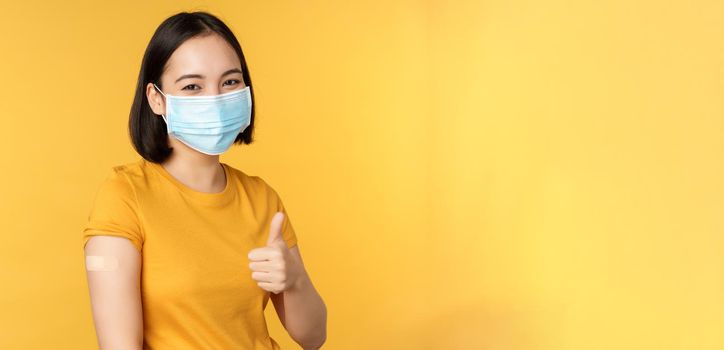 Vaccination from covid and health concept. Happy asian girl showing thumbs up, wearing medical mask, band aid on shoulder, got coronavirus vaccine shot, yellow background.