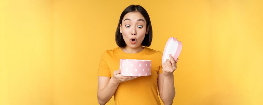 Portrait of excited asian woman, open gift box with surprised happy face, standing over yellow background.