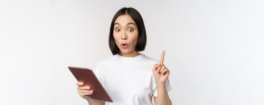 Enthusiastic asian woman with tablet, raising finger and looking amazed, pointing up, standing over white background.