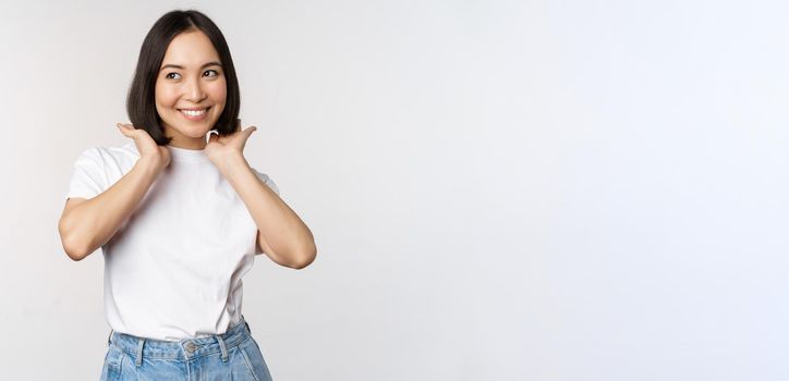 Portrait of cute, beautiful asian woman touching her new short haircut, showing hairstyle, smiling happy at camera, standing over white background.