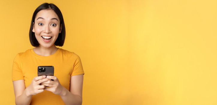 Happy asian girl smiling, standing with black mobile phone, standing against yellow background. Copy space