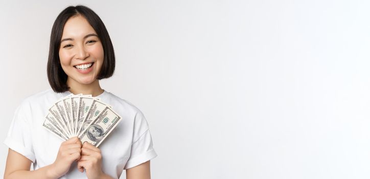 Portrait of smiling asian woman holding dollars money, concept of microcredit, finance and cash, standing over white background.