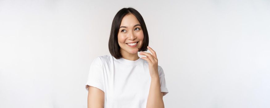 Portrait of cute coquettish woman laughing and smiling, looking aside thoughtful, thinking or imaging smth, standing in white t-shirt over studio background.