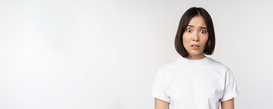 Image of shocked and frustrated asian woman, looking alarmed at camera, cant understand, puzzled, standing over white background.