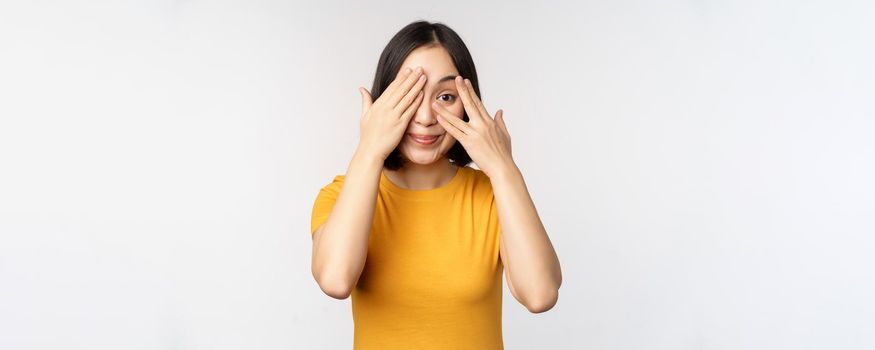 Cute korean female model, cover eyes with hands, peeking through fingers curious, watching smth, standing in yellow tshirt over white background.