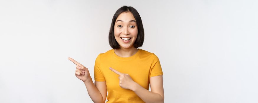 Portrait of smiling asian brunette girl in yellow tshirt, pointing fingers left, showing copy space, promo deal, demonstrating banner, standing over white background.