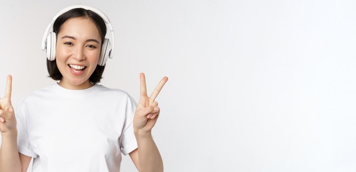 Happy asian woman wearing headphones and smiling, showing peace v-sign, listening music, standing in tshirt over white background.
