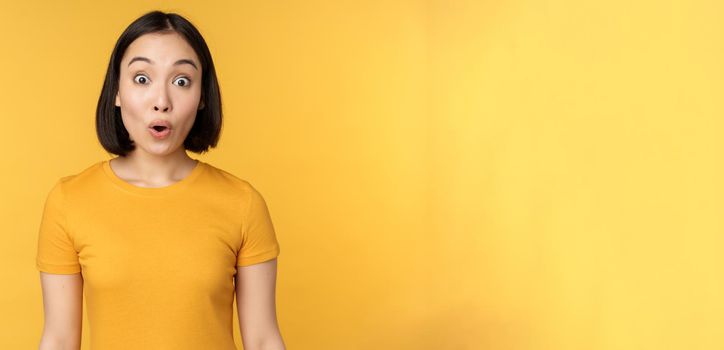 Image of asian girl looking surprised, reacting amazed, raising eyebrows impressed, standing over yellow background.