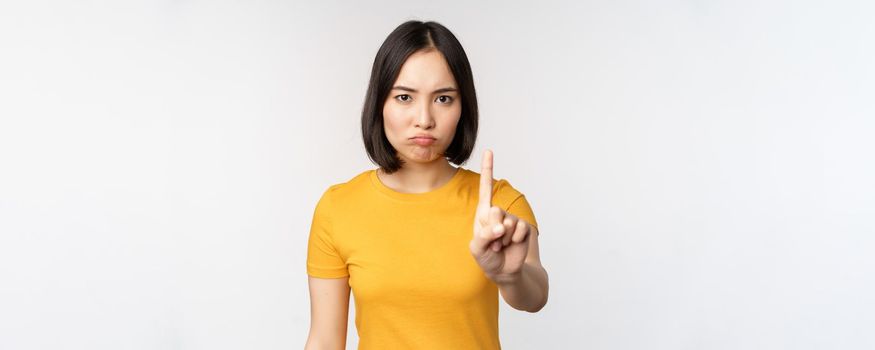 Portrait of asian woman looking serious and angry, showing stop prohibit gesture, taboo sign, forbidding smth, standing in yellow tshirt over white background.