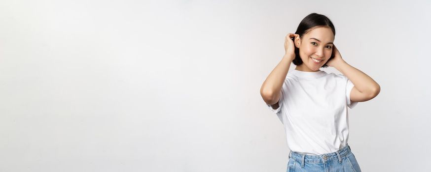 Portrait of cute, beautiful asian woman touching her new short haircut, showing hairstyle, smiling happy at camera, standing over white background.