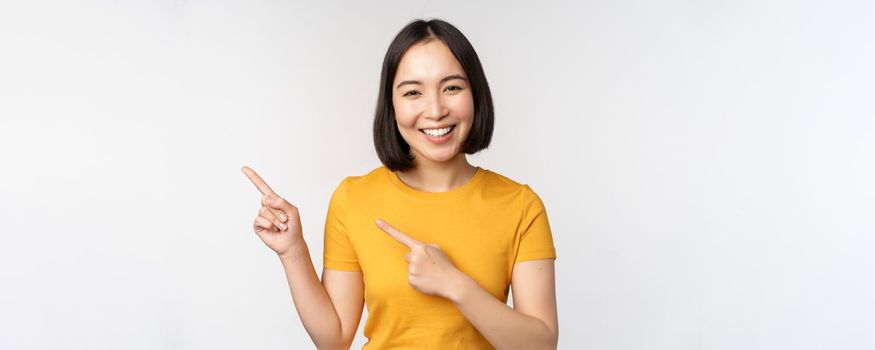 Portrait of smiling asian brunette girl in yellow tshirt, pointing fingers left, showing copy space, promo deal, demonstrating banner, standing over white background.