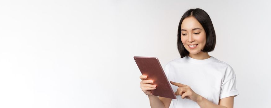 Asian girl using digital tablet, working with gadget, shopping online, standing over white background.