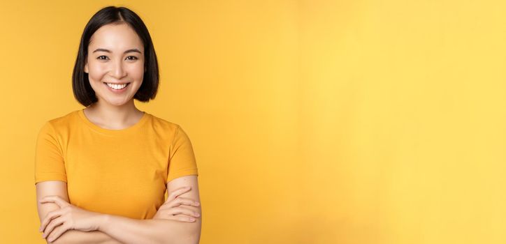 Confident asian girl cross arms on chest, smiling and looking assertive, standing over yellow background.
