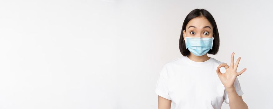 Covid-19, healthcare and medical concept. Impressed asian woman in medical mask, looking amazed and showing ok, okay sign, standing over white background.