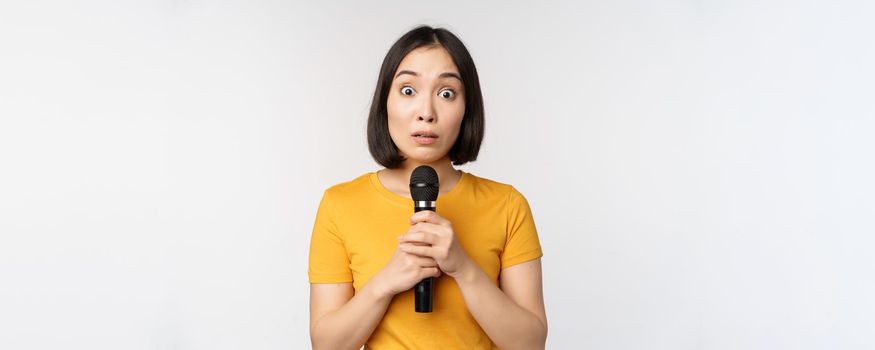 Modest asian girl holding microphone, scared talking in public, standing against white background. Copy space