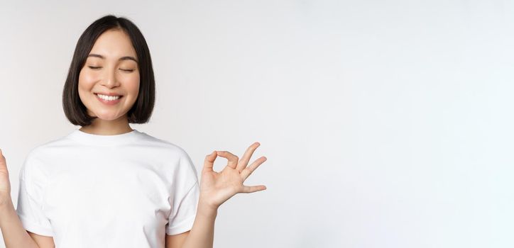 Portrait of young asian woman meditating, smiling pleased and practice yoga, close eyes and meditate, breathing calm, standing over white background.