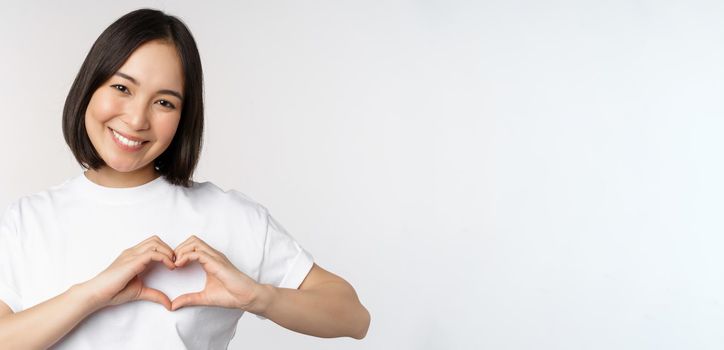 Lovely asian woman smiling, showing heart sign, express tenderness and affection, standing over white background.