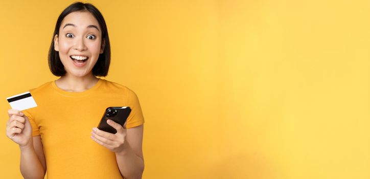 Online shopping. Cheerful asian girl holding credit card and smartphone, paying, order with mobile phone, standing over yellow background.