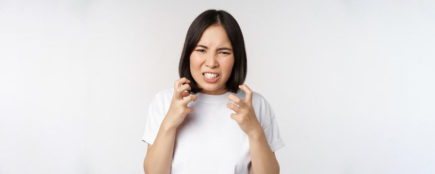 Angry asian woman cursing, looking outraged and annoyed, clench teeth and frowning furious, standing over white background.