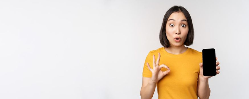 Excited asian girl showing mobile phone screen, okay sign, recommending smartphone app, standing in yellow tshirt over white background.