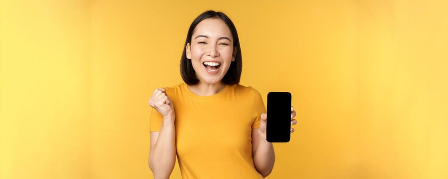 Excited asian woman, showing smartphone app and triumphing, celebrating on mobile phone, standing over yellow background. Copy space