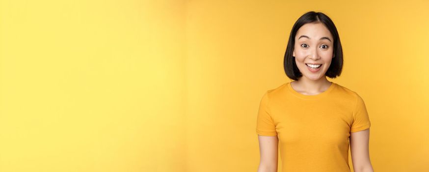 Image of asian girl looking surprised, reacting amazed, raising eyebrows impressed, standing over yellow background.