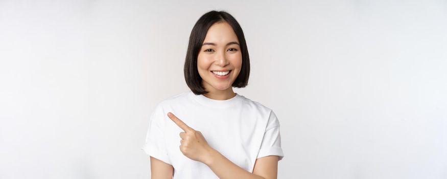 Close up of young japanese woman smiling, pointing finger left at copy space, showing announcement or advertisment banner, standing over white background.