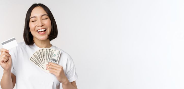 Portrait of asian woman smiling, holding credit card and money cash, dollars, standing in tshirt over white background.