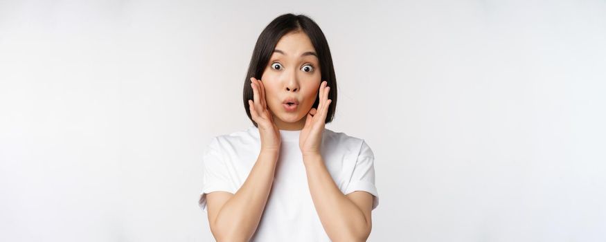 Close up portrait of asian girl looking surprised, wow face, reacting amazed at smth, standing in white tshirt over studio background, isolated.